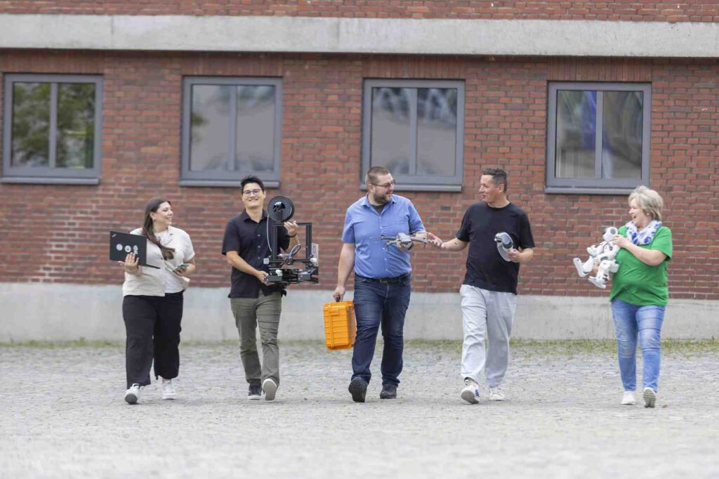 Eine Gruppe von Menschen traegt digitale Geraete vor einem Backsteingebaeude.