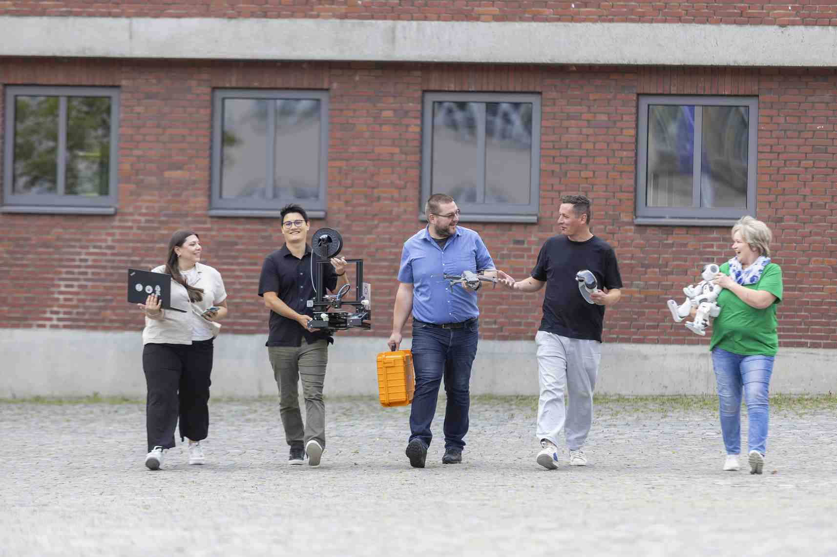 Eine Gruppe von Menschen traegt digitale Geraete vor einem Backsteingebaeude.