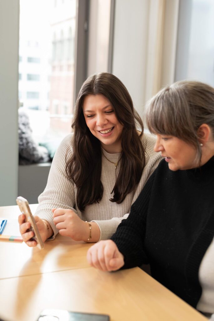2 Frauen sitzen am Tisch und schauen gemeinsam auf ein Smartphone.