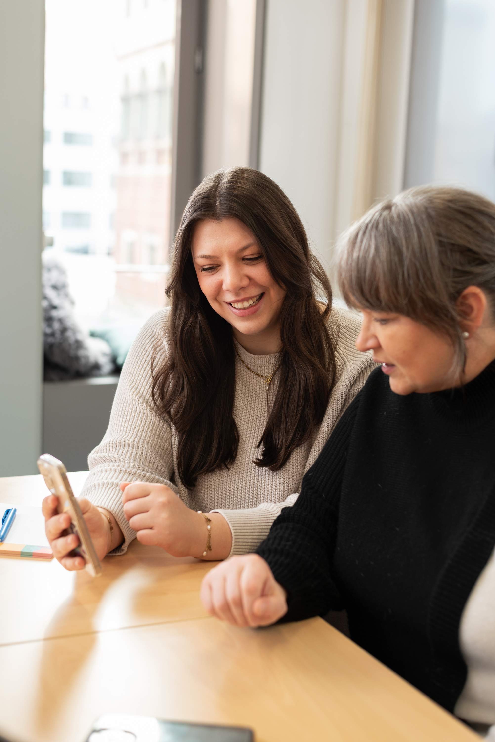 2 Frauen sitzen am Tisch und schauen gemeinsam auf ein Smartphone.