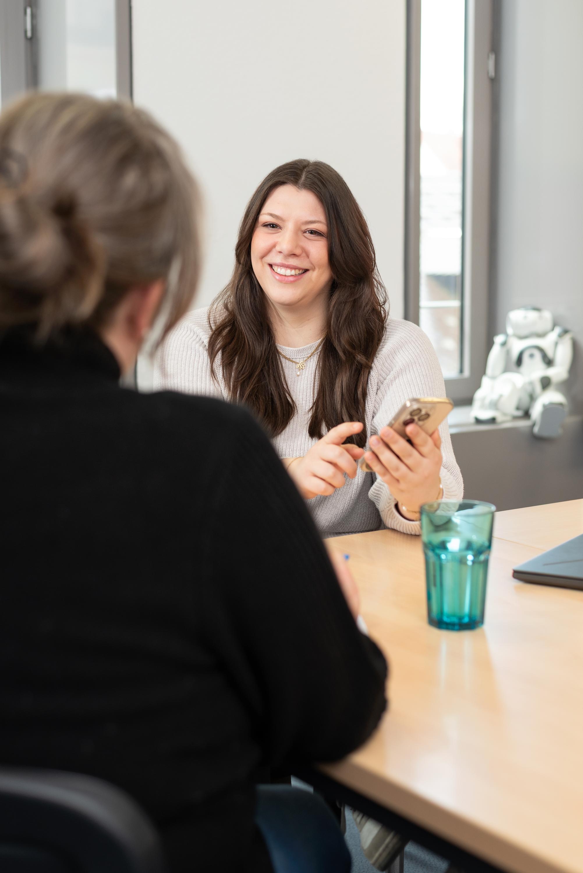 Zwei Personen bei einer Beratung im Digitalisierungszentrum. Eine Frau haelt ein Smartphone in der Hand. Blau, Braun, Grau
