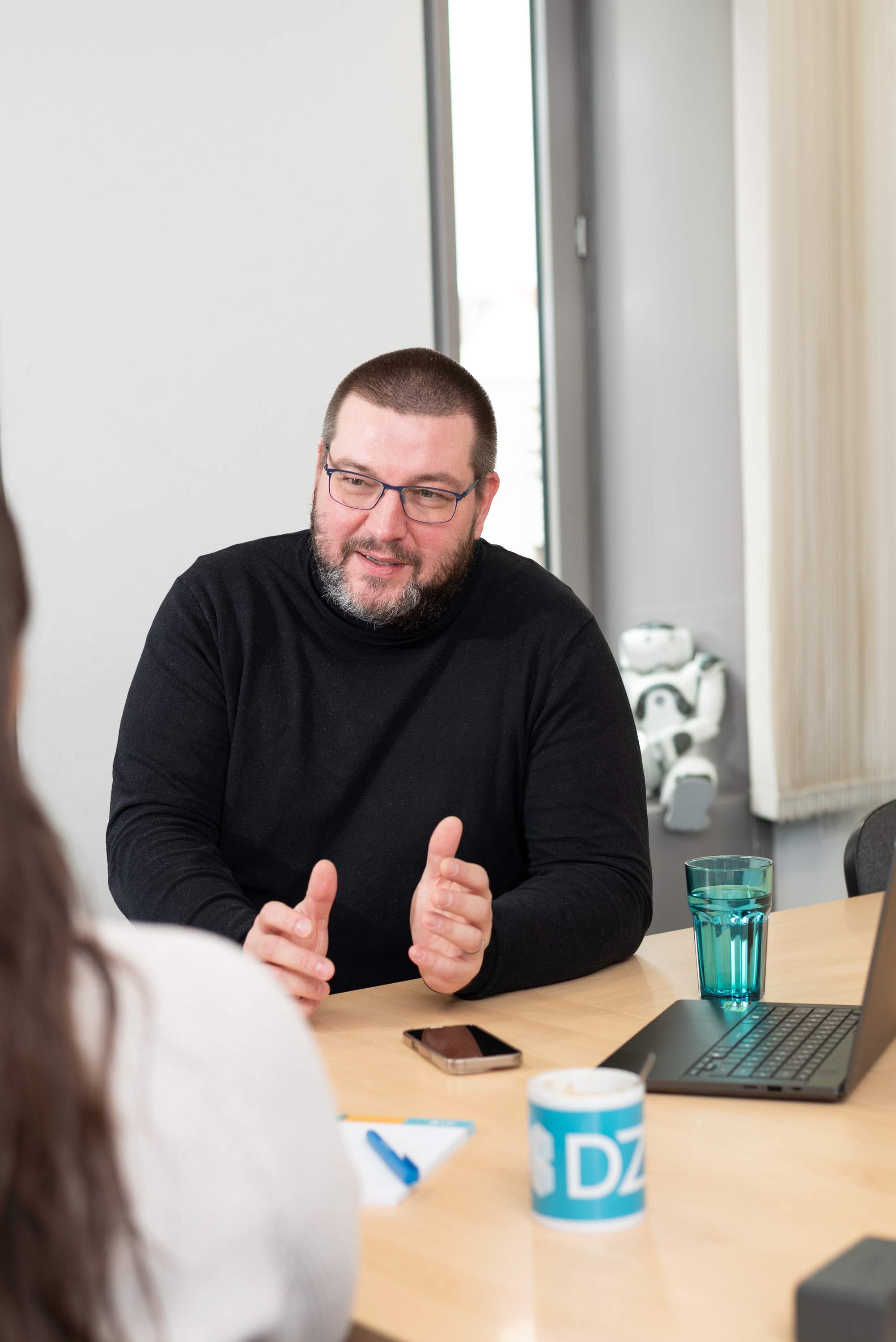 Ein Mann in einer Beratungssituation sitzt an einem Tisch. Vor ihm steht ein Wasserglas und liegt ein Smartphone.
