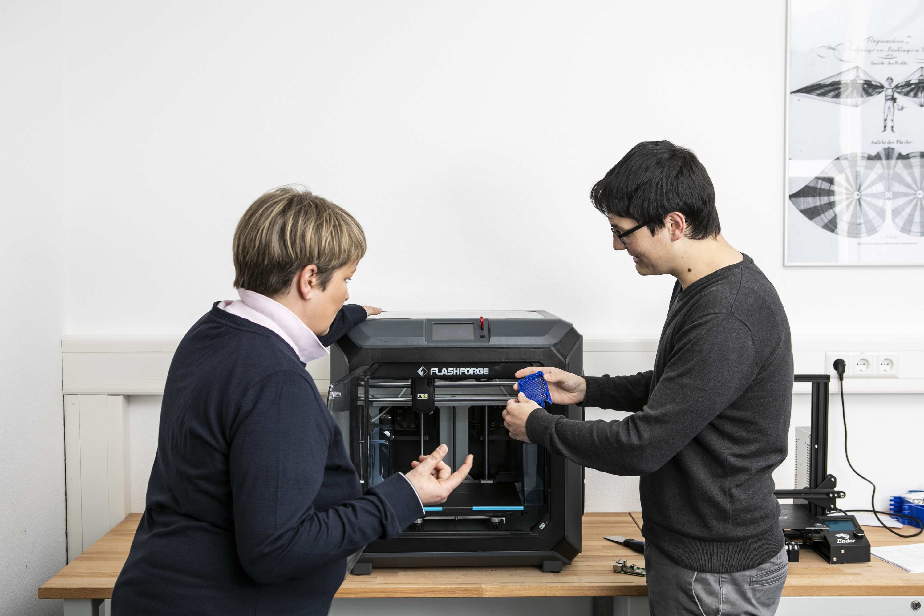 Zwei Personen arbeiten gemeinsam an einem 3D-Drucker im Digitalisierungszentrum Ulm. Grau, Schwarz, Blau
