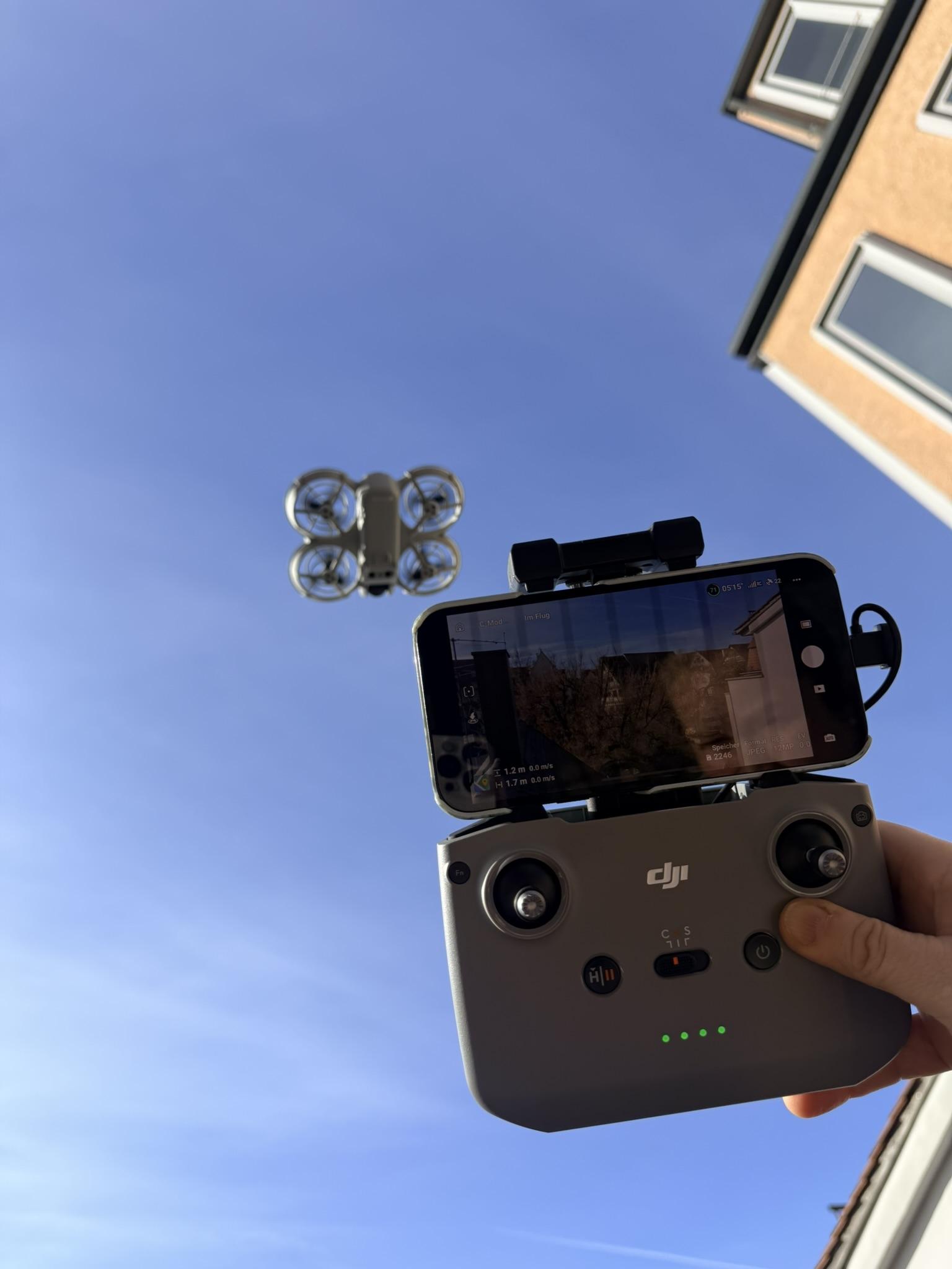 Steuerung einer Drohne mit Fernbedienung und Smartphone im Himmel ueber einem Gebaeude.