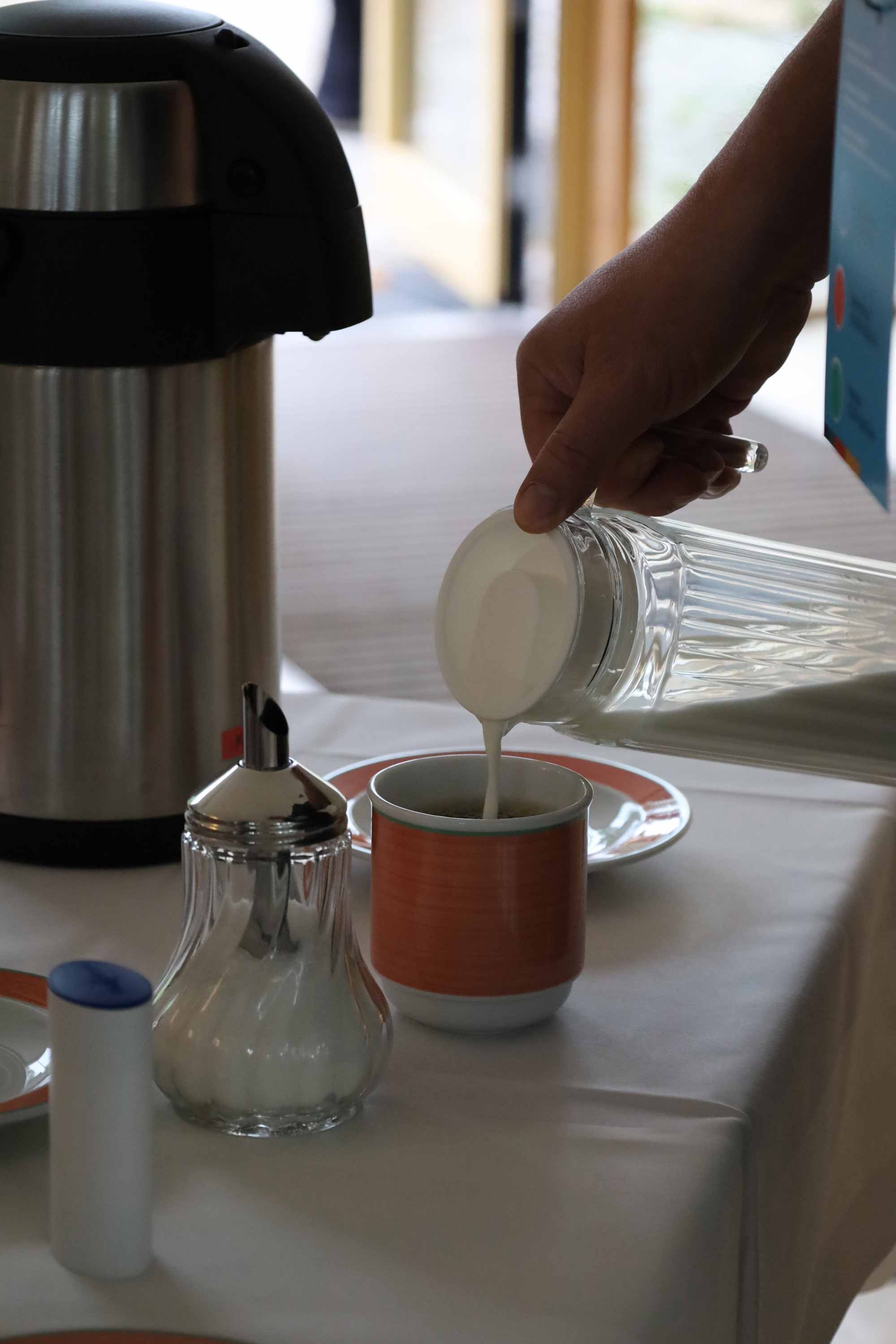 Eine Hand giesst Milch aus einem Glaskrug in eine Tasse neben Thermoskanne und Zuckerstreuer.