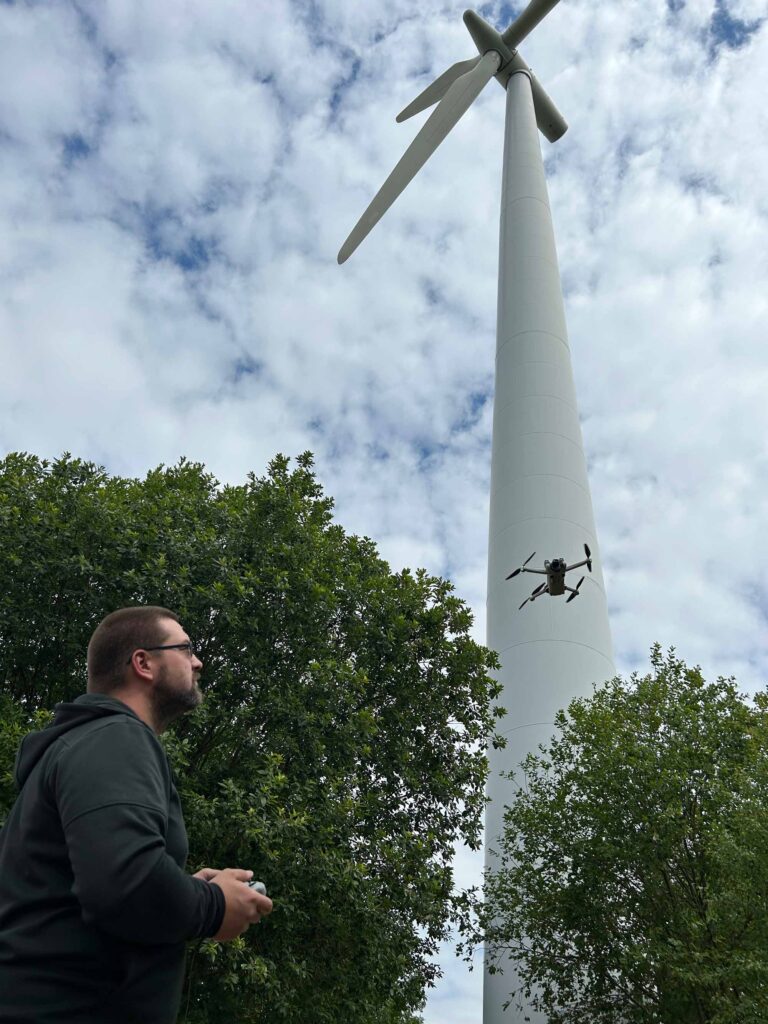 Person steuert eine Drohne vor einer Windkraftanlage unter bewoelktem Himmel.