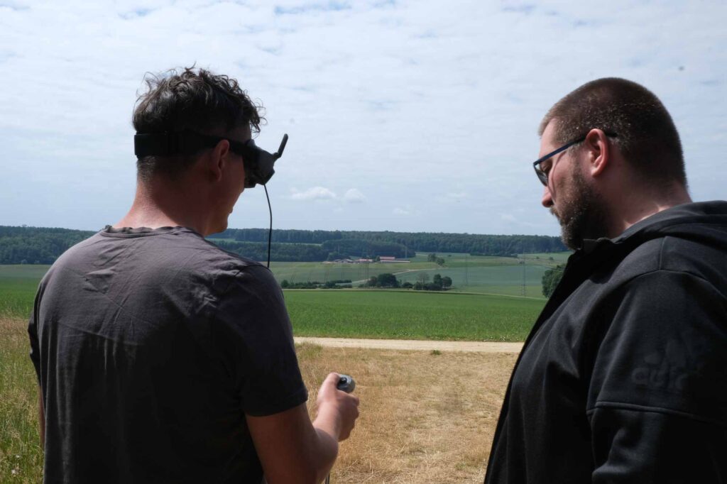 Zwei Personen steuern eine Drohne in laendlicher Umgebung, eine davon traegt eine Videobrille.