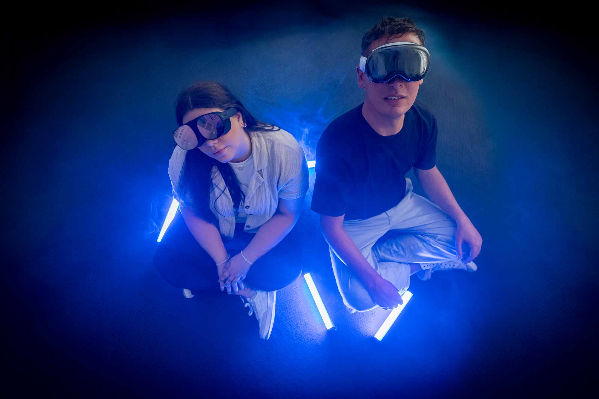 Zwei Personen mit unterschiedlichen Virtual Reality Brillen sitzen zwischen blau leuchtenden Lichtstaeben in einem dunklen Raum.