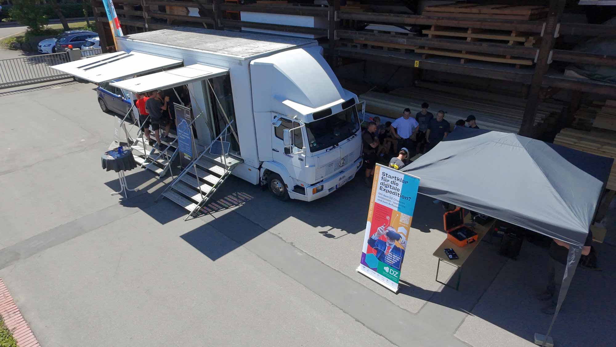 Mobiles Digitalisierungslabor mit ausgefahrenen Treppen und Vordach steht neben einem Informationsstand und einer Gruppe von Personen.