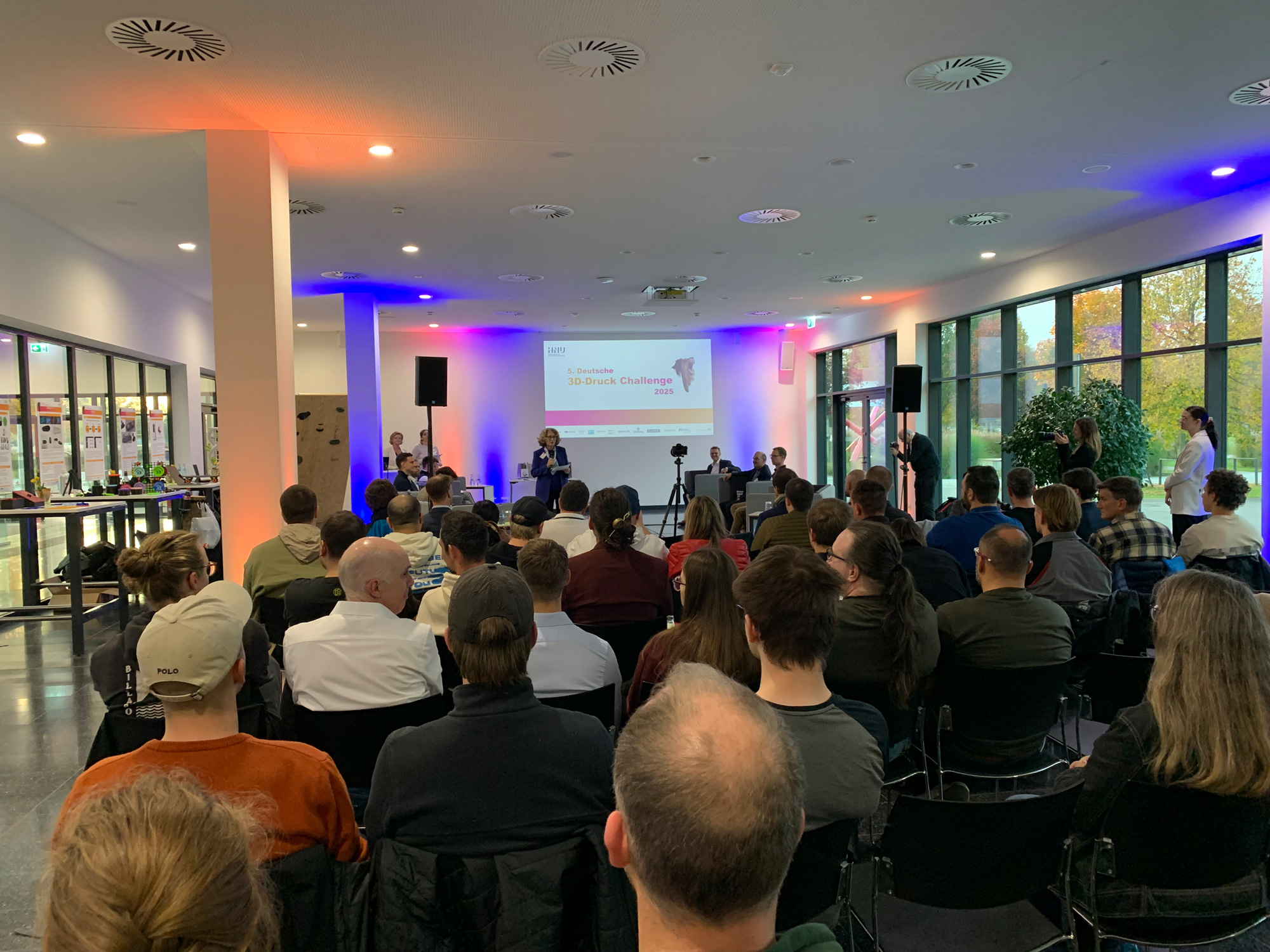 Publikum sitzt vor einer Buehne bei der Deutschen 3D Druck Challenge 2025 mit Praesentation und ausgestellten Projekten.