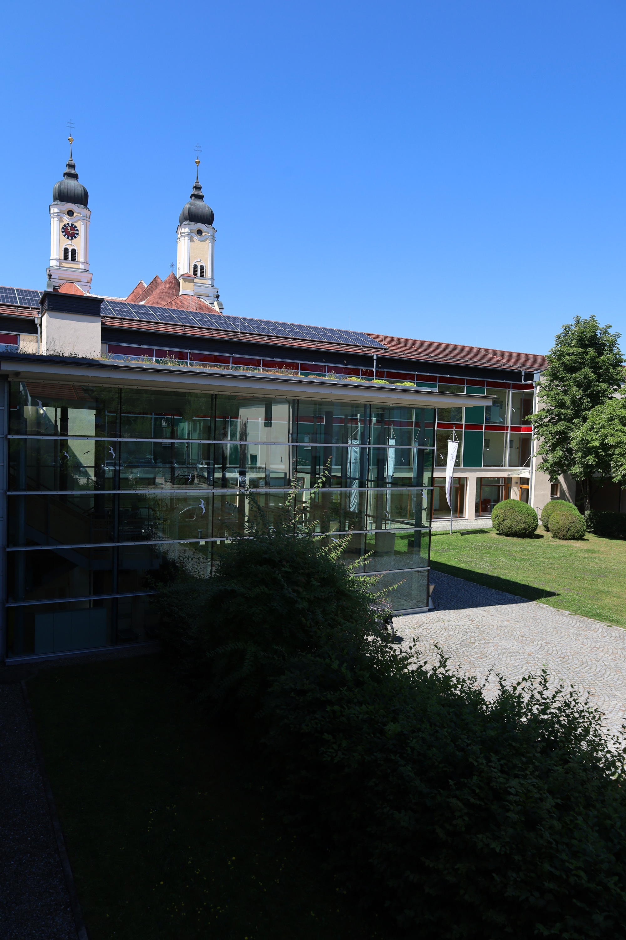 Moderner Gebaeudekomplex mit Glasfassade und Solaranlage vor zwei Kirchtuermen unter blauem Himmel.