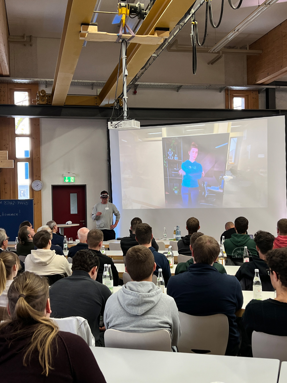 Vortragender mit VR Brille steht vor einem Publikum und einer Leinwand mit einer virtuellen Person.
