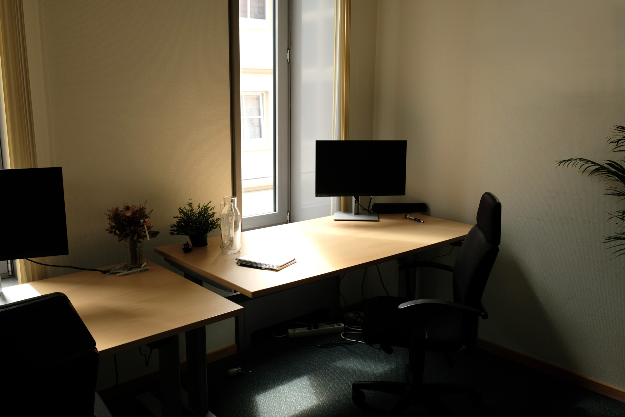 Zwei Arbeitsplaetze mit Monitoren und Buerostuehlen stehen an einem sonnigen Fenster.