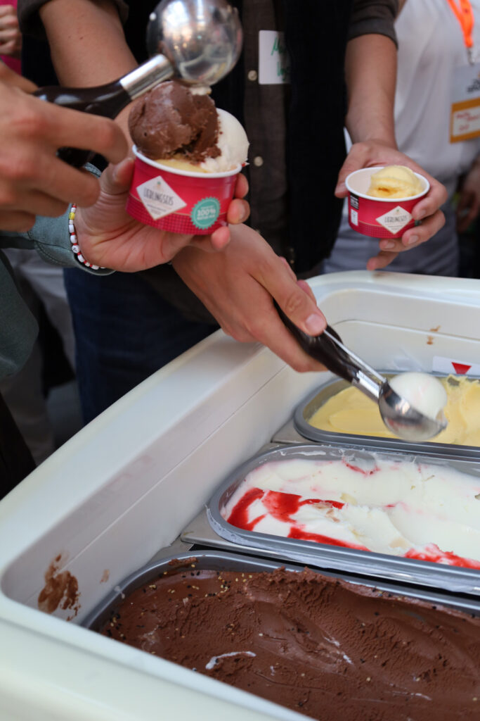 Mehrere Haende fuellen verschiedene Eissorten aus einer Eistheke in kleine Becher.