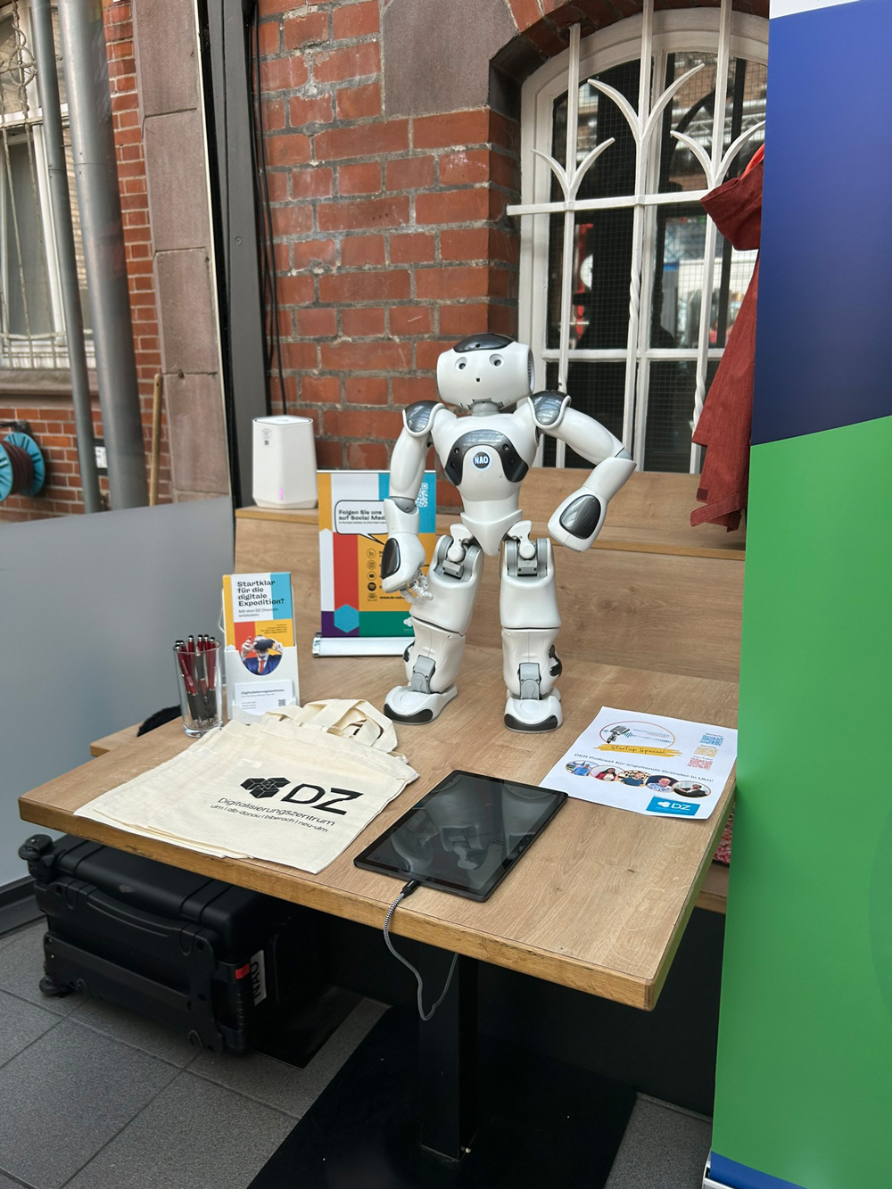 Humanoider Roboter steht auf einem Infotisch mit Tablet, Stofftaschen und Informationsmaterial.