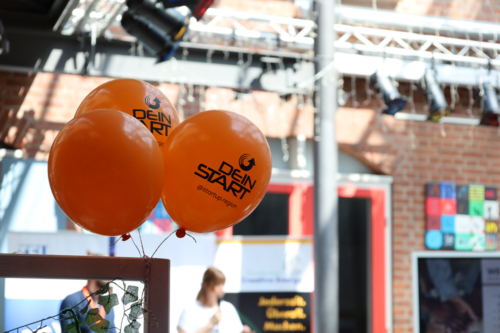 Drei orange Luftballons mit der Aufschrift Dein Start haengen in einem Veranstaltungsraum.