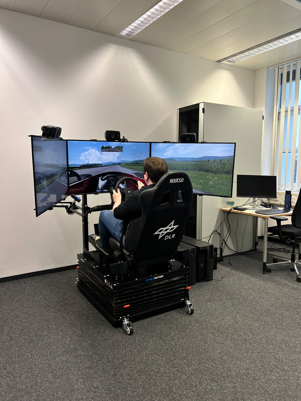 Ein Mann sitzt in einem Fahrsimulator mit Lenkrad und drei Bildschirmen, die eine virtuelle Landstrasse zeigen.