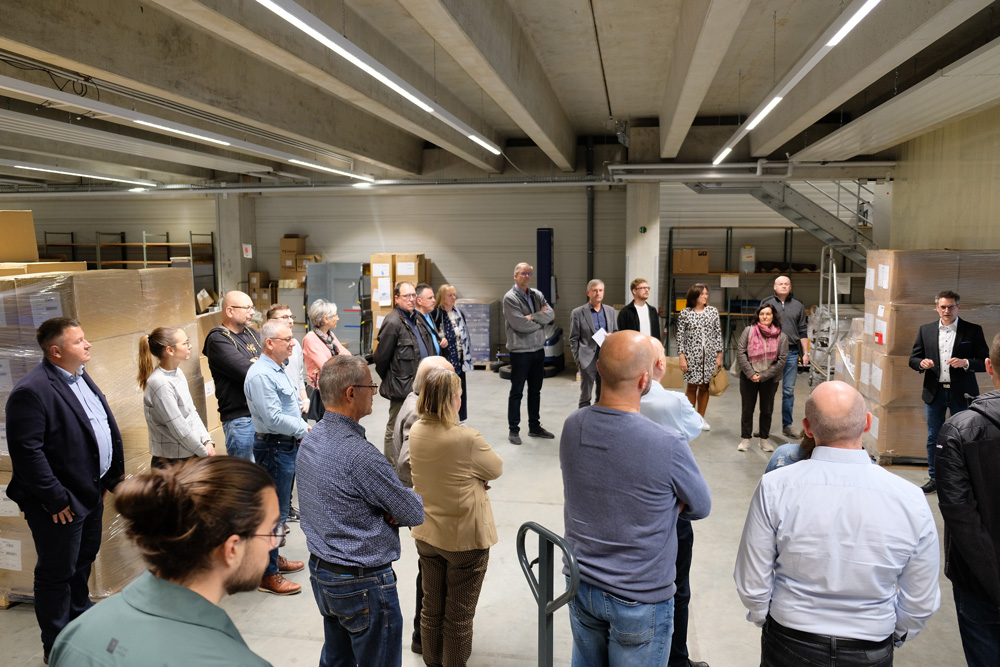 Eine Gruppe von Menschen steht in einer Lagerhalle zwischen Paletten und Kartons und hoert einem Vortragenden zu.
