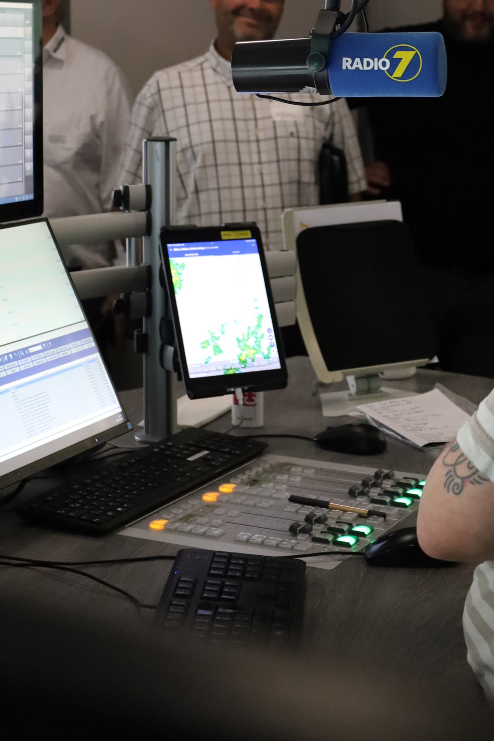 Radio 7 Mikrofon ueber einem Mischpult und mehreren Monitoren mit Personen im Hintergrund eines Radiostudios.