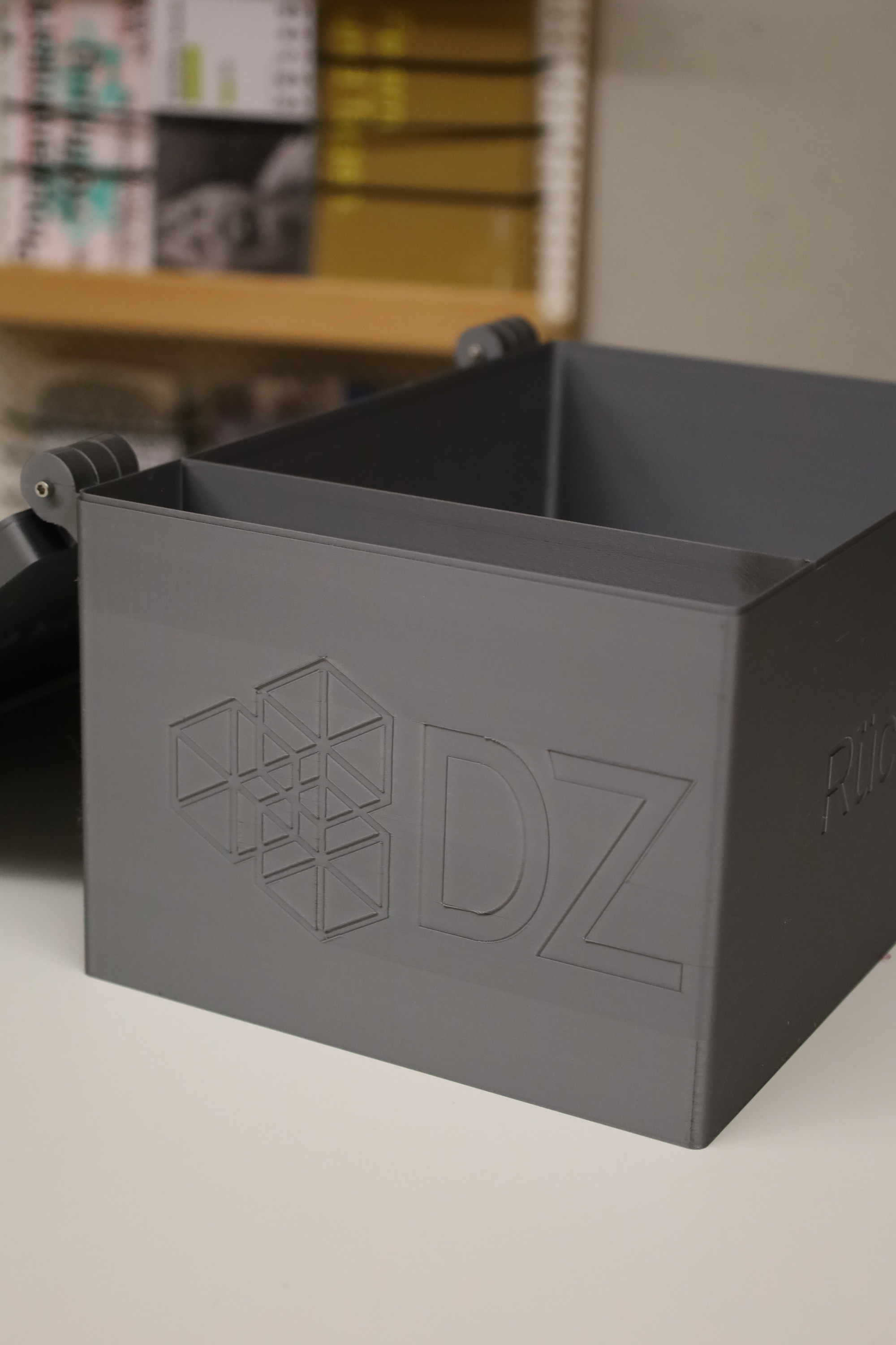 Graue 3D gedruckte Box mit eingeprägtem DZ Logo auf einem Tisch.