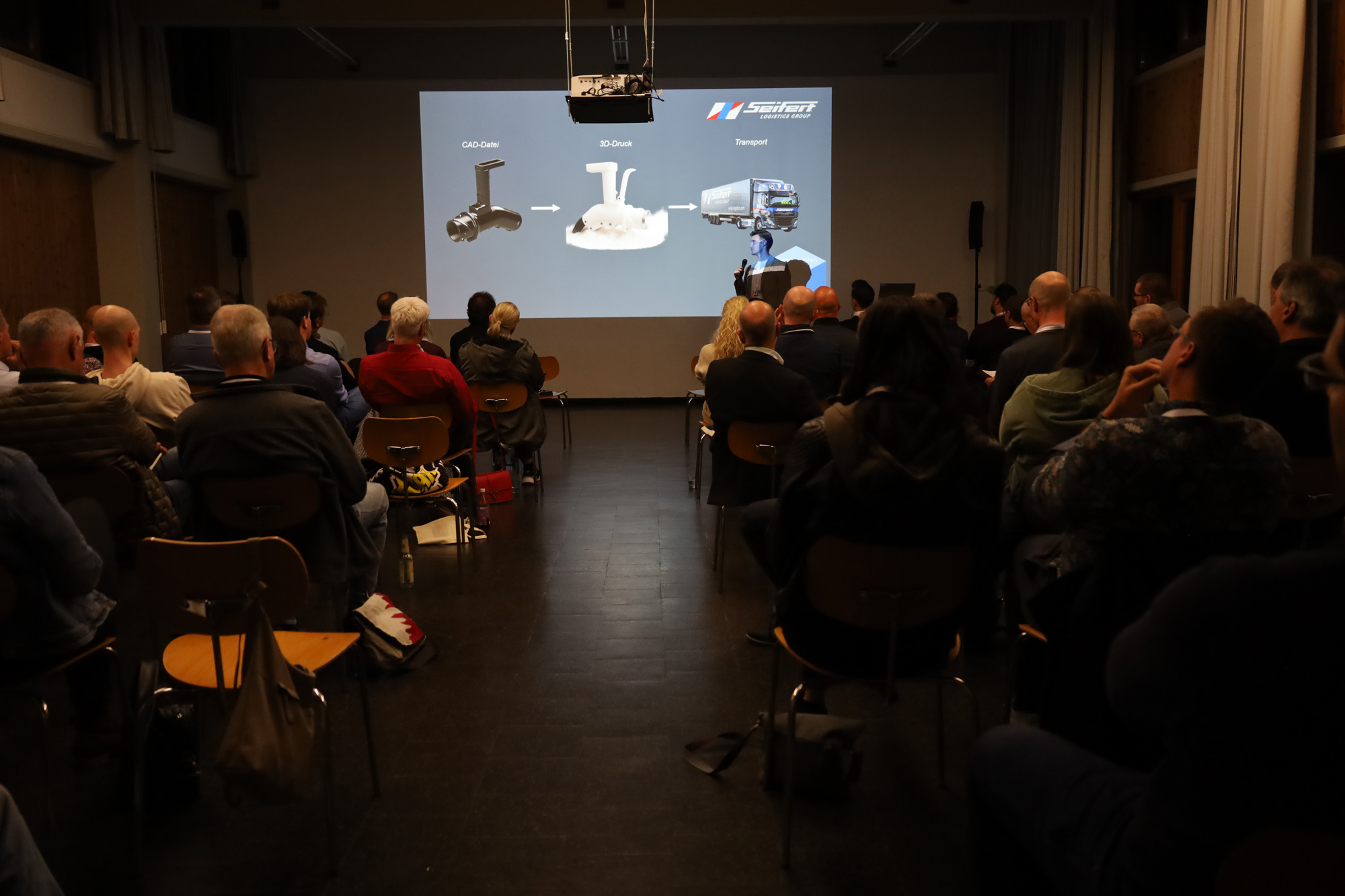 Vortragender mit Mikrofon praesentiert vor einem grossen Publikum eine Folie zu CAD Datei, 3D Druck und Transport.