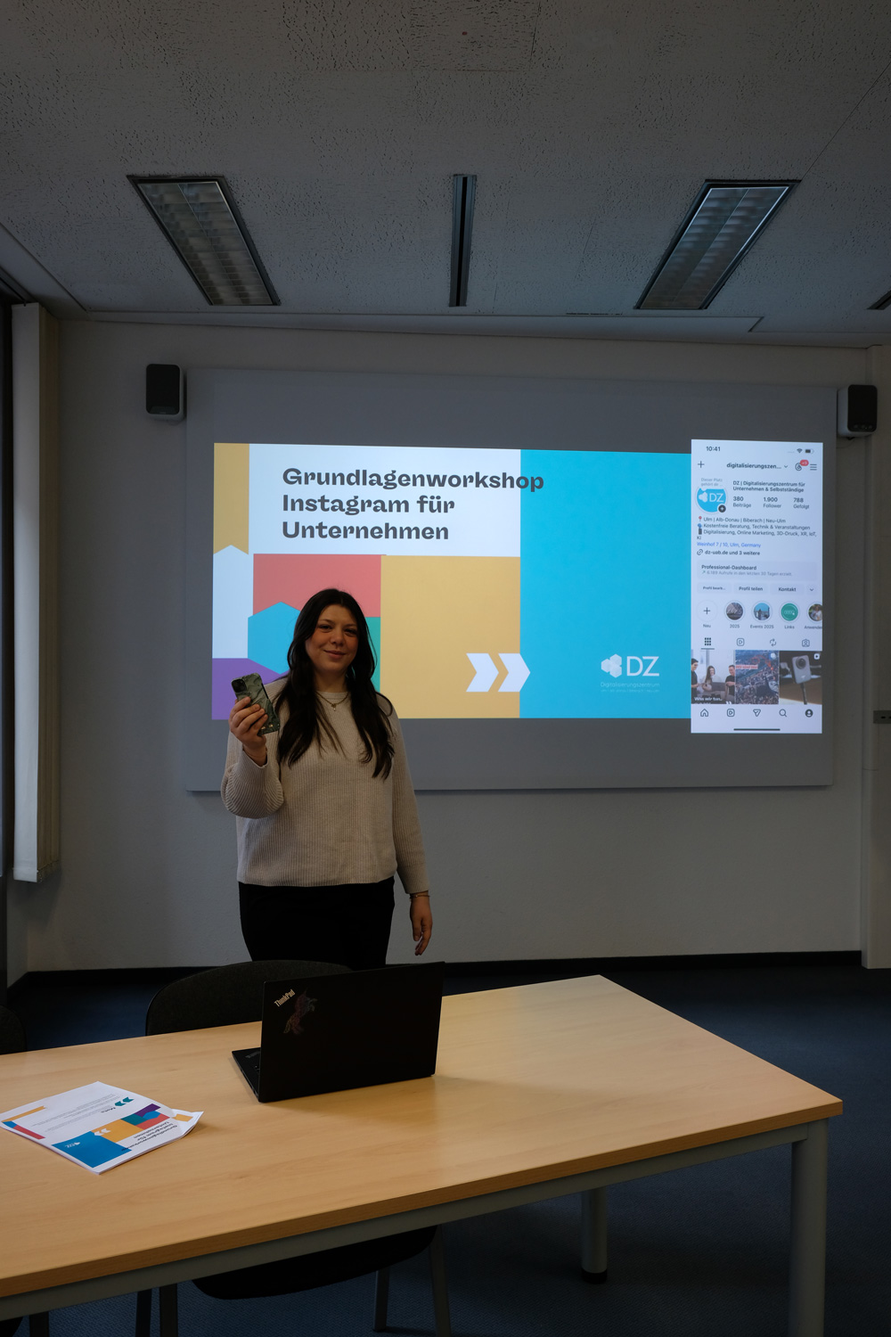 Eine Frau steht mit Smartphone vor einer Praesentationsfolie zum Grundlagenworkshop Instagram fuer Unternehmen.