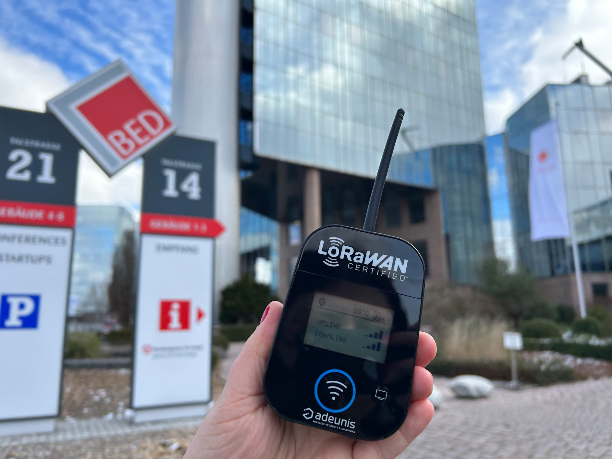 Hand haelt ein LoRaWAN Messgeraet vor dem BED in Ehingen mit Wegweisern im Hintergrund.