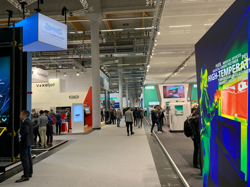 Besucher gehen durch eine Messehalle mit zahlreichen Ausstellungsstaenden und grossen digitalen Praesentationsflaechen.