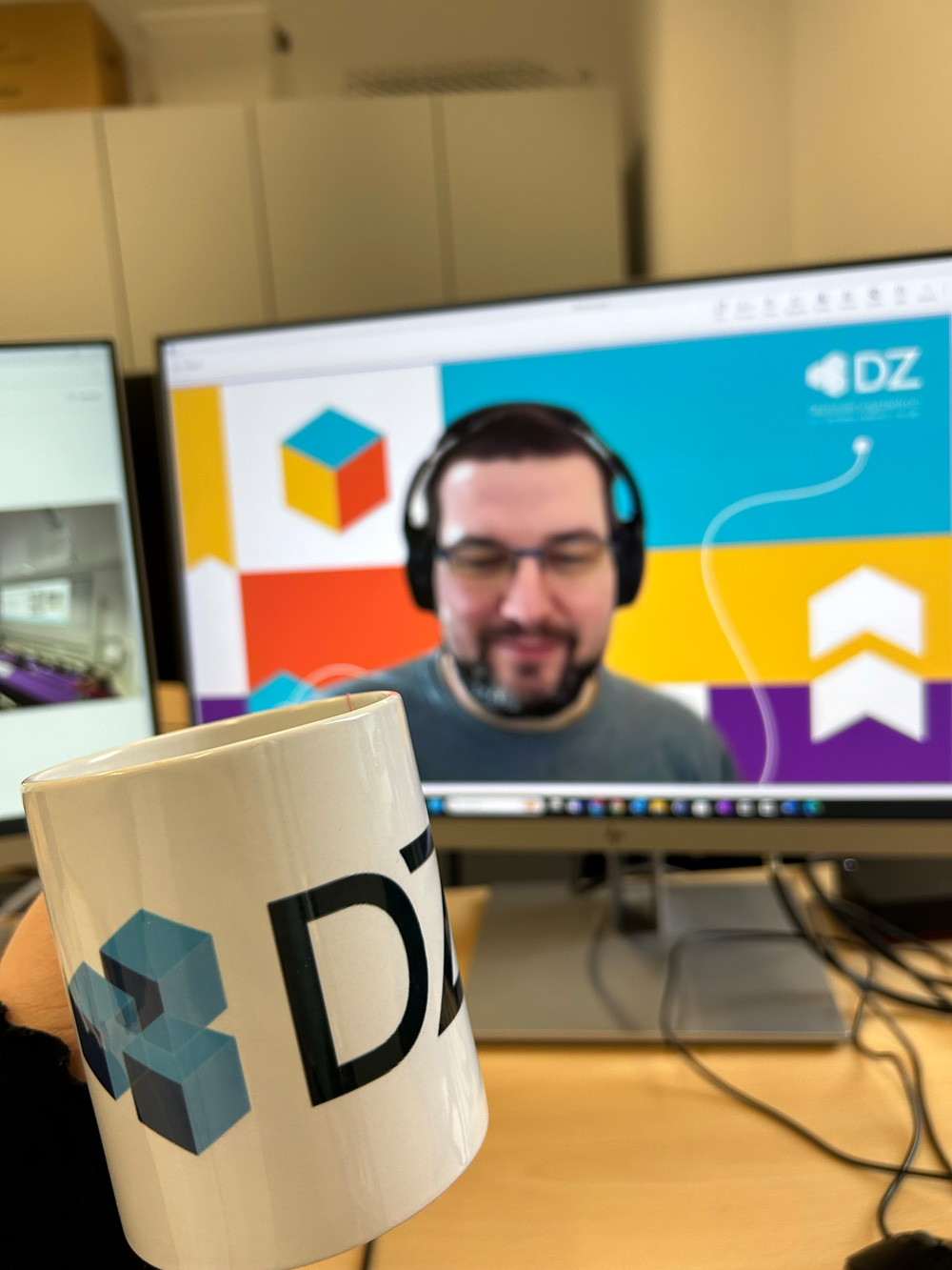 Tasse mit DZ Logo vor einem Bildschirm mit einer Person mit Kopfhoerern in einer Videokonferenz.
