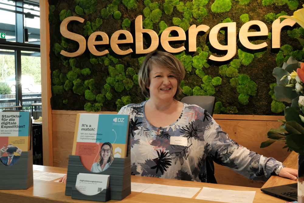 Eine laechelnde Frau sitzt an einem Informationsstand vor einer gruenen Mooswand mit dem Schriftzug Seeberger.