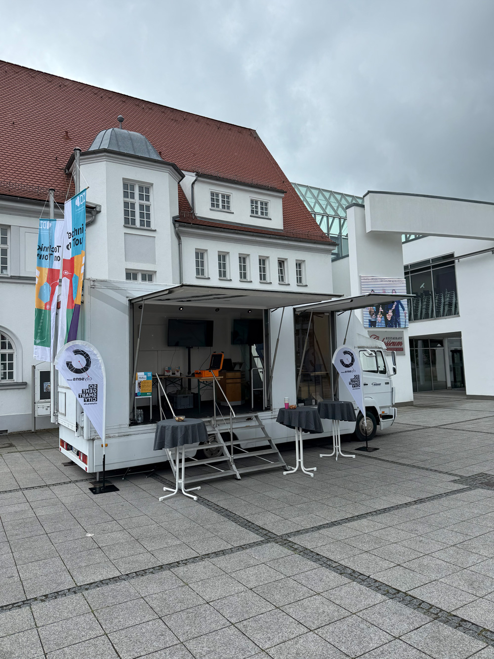 Mobiler Showtruck mit geoeffneter Ausstellungsflaeche und Fahnen steht vor einem Veranstaltungsgebaeude.