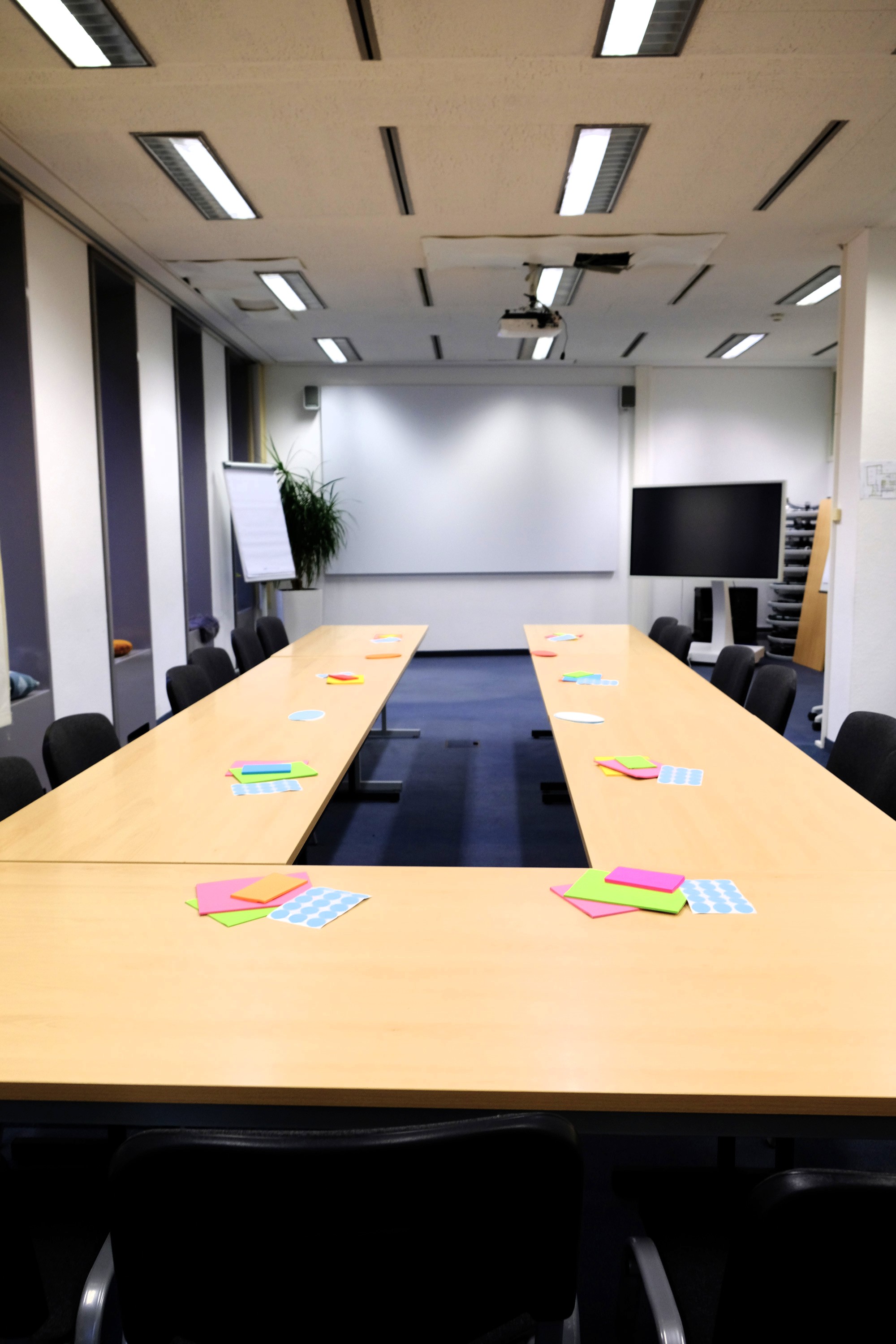 Leerer Workshopraum mit U foermig angeordneten Tischen, bunten Haftnotizen, Stuehlen, Flipchart und Bildschirm.