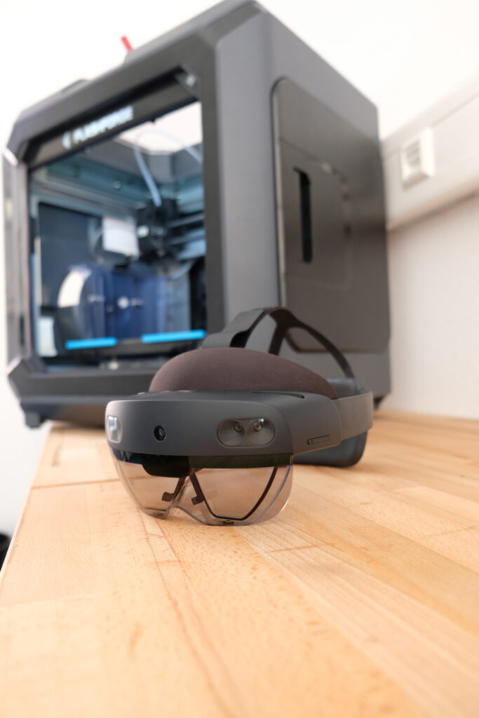 Augmented Reality Brille liegt auf einem Holztisch vor einem geschlossenen 3D Drucker.