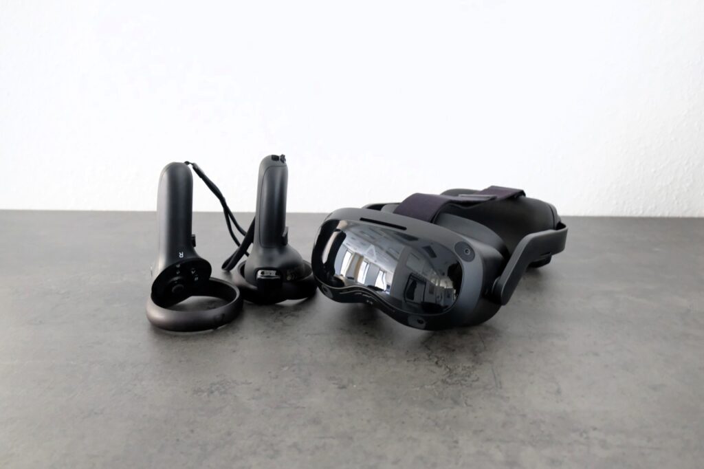 Schwarze VR Brille mit zwei Controllern liegt auf einer grauen Tischflaeche.