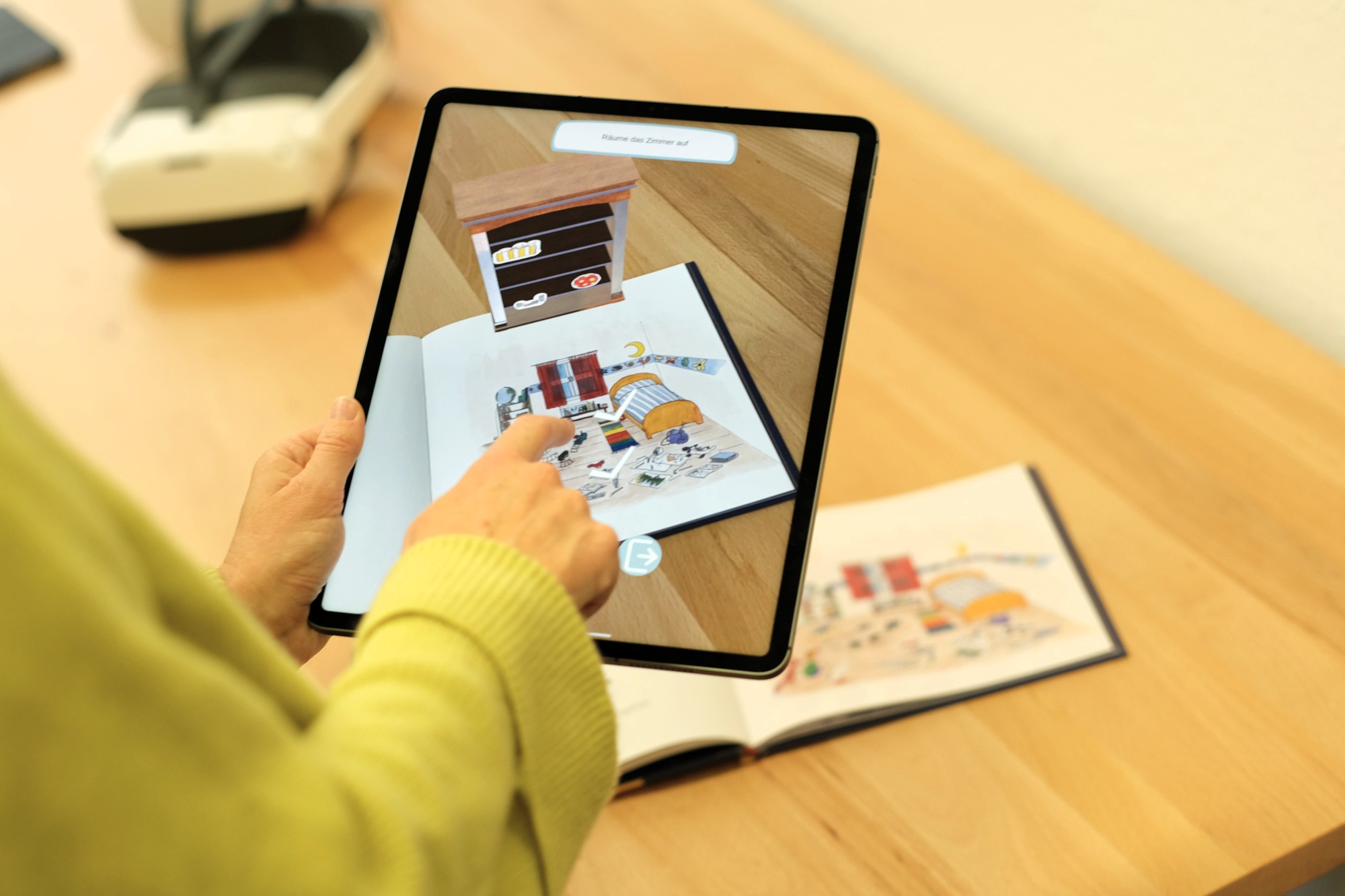 Person bedient ein Tablet mit einer interaktiven Darstellung eines Kinderzimmers ueber einem aufgeschlagenen Bilderbuch.