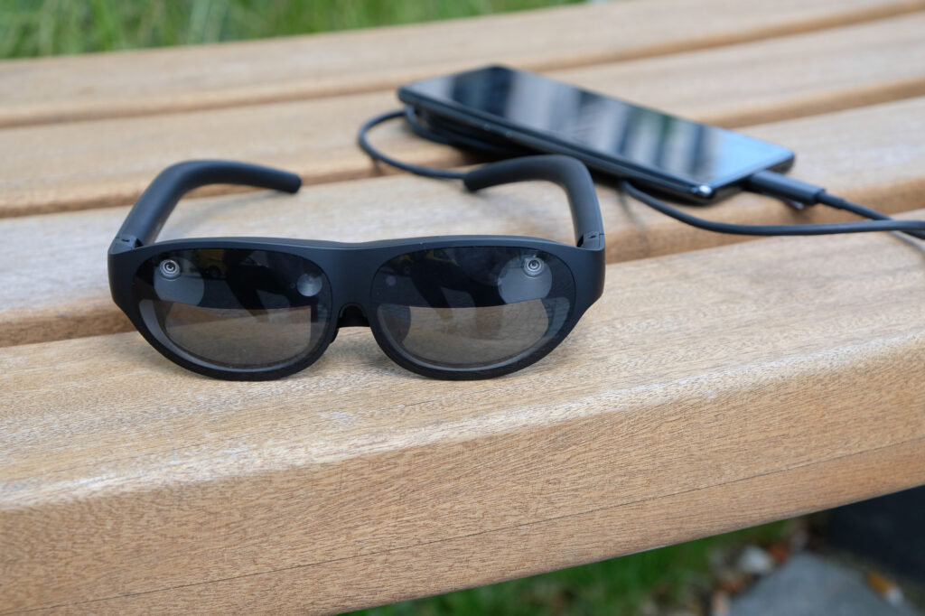 Schwarze Datenbrille und ein per Kabel verbundenes Smartphone liegen auf einer Holzbank. Weisse VR Brille mit zwei Controllern liegt auf einem hellen Holztisch.