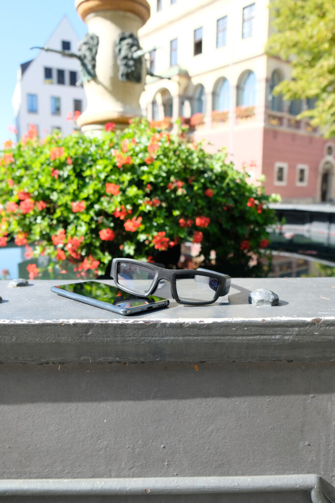 Schwarze Smart Brille und Smartphone liegen auf einer Mauer vor roten Blumen und historischen Gebaeuden.
