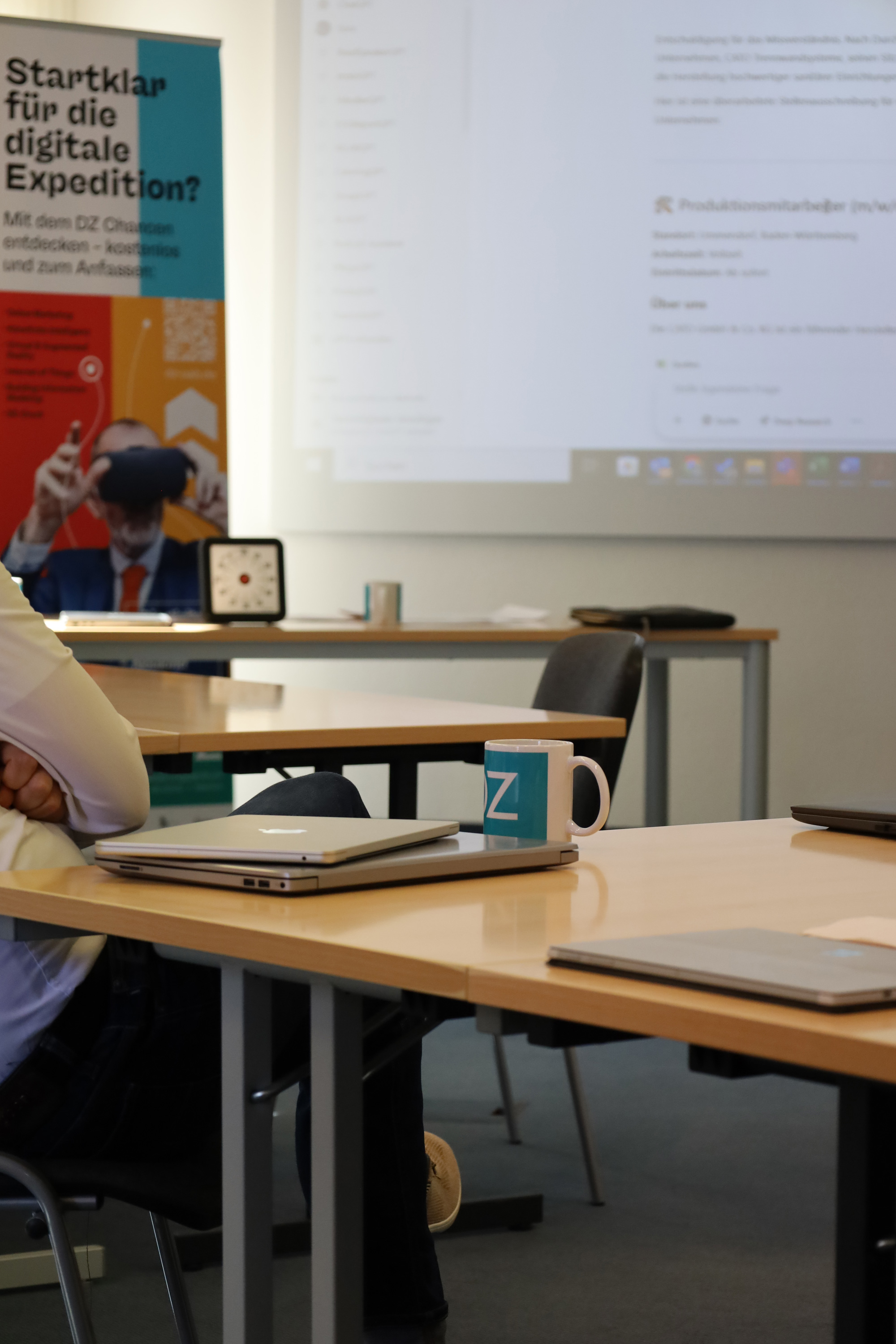 Seminarraum mit Laptops auf Tischen, einer DZ Tasse und einer projizierten Webseite im Hintergrund.